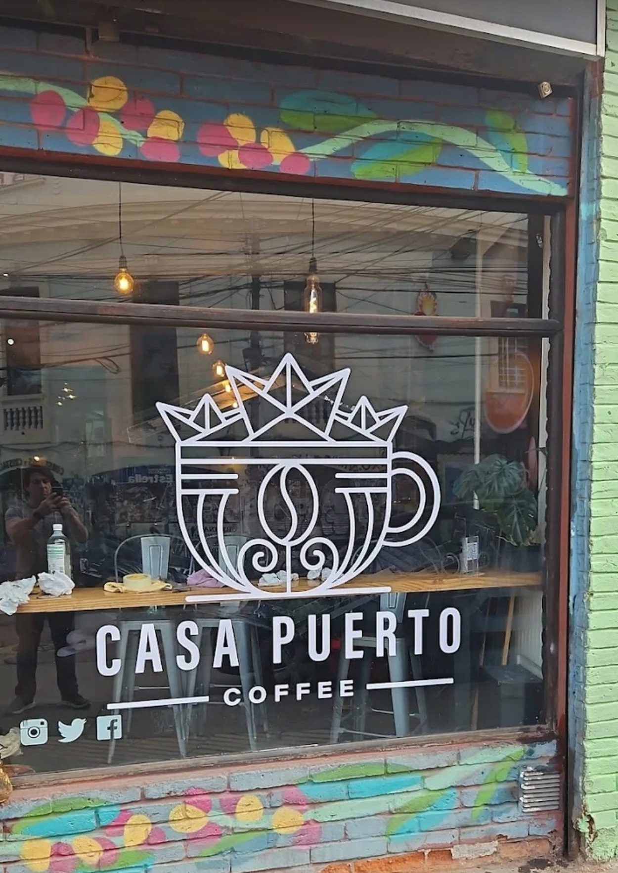 Vitrina de Casa Puerto Coffee en Condell 1154: muro de ladrillo pintado con un mural de flores y la taza coronada por barquitos de papel aplicada en vinilo blanco sobre el vidrio