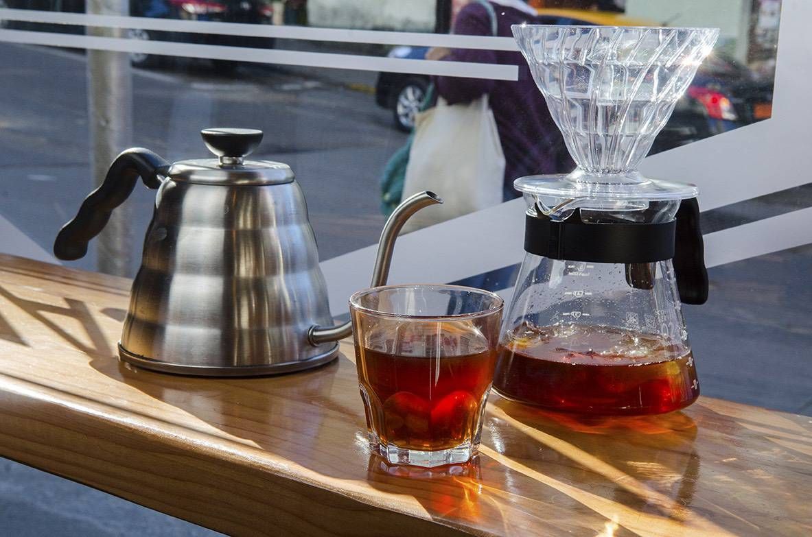 Cafetera de cuello de cisne, un vaso de café filtrado y una jarra sobre la barra de madera junto al ventanal, con la calle y la gente pasando detrás del vidrio
